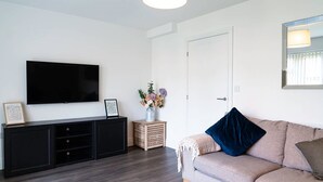 A modern living room with a flat-screen TV mounted on a black cabinet, a beige sofa with a blue pillow, and a small side table with a vase of flowers. - Host Stay Hewitt Close (Peterborough)