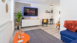 A modern living room with a flat-screen TV mounted on the wall, a sofa, a small table with a bottle and glasses, and a potted plant. - Host Stay Davies Close (Market Harborough)