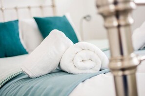 Un letto rifatto con cura, un asciugamano bianco e un asciugamano arrotolato, cuscini color acquamarina e una testiera in metallo.