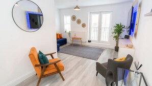 A modern living room with a blue sofa, orange armchair, and a glass coffee table. - Host Stay Davies Close (Market Harborough)