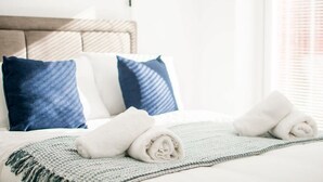A neatly made bed with rolled white towels, a grey knitted blanket, and two pillows (one blue and one white). - Host Stay Davies Close (Market Harborough)
