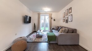Ein Wohnzimmer mit grauem Sofa, einem kleinen Couchtisch, einem an der Wand befestigten Fernseher und einem Fenster mit Vorhängen.
