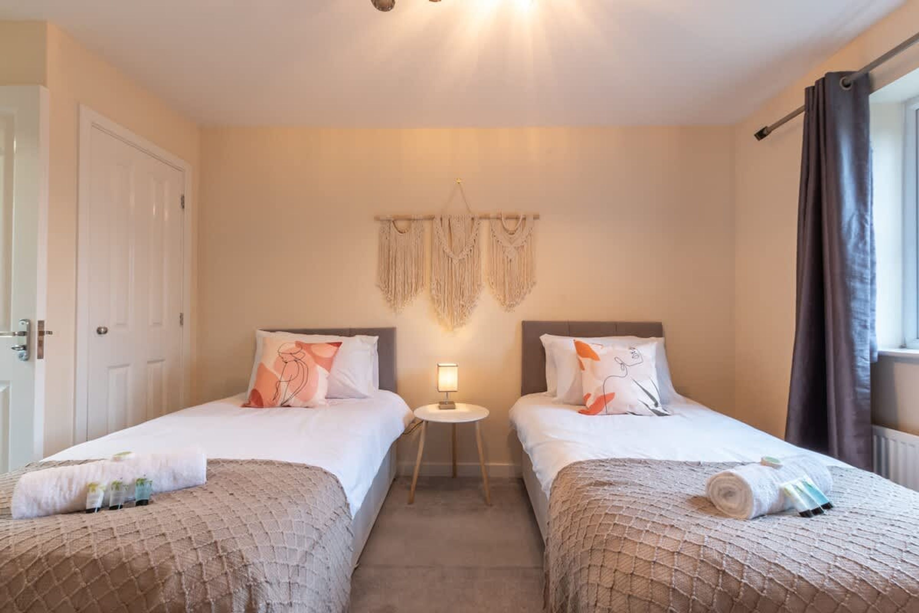 A hotel room with two single beds, a small bedside table with a lamp, a wall-mounted macrame decoration, and a window with curtains.