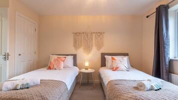 A hotel room with two single beds, a small bedside table with a lamp, a wall-mounted macrame decoration, and a window with curtains.