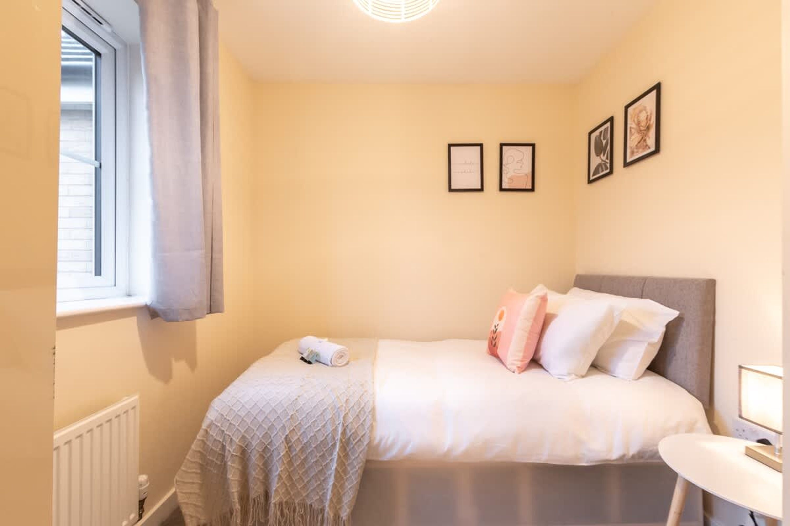 Un dormitorio con cama, mesita de noche, ventana con cortinas e imágenes enmarcadas en la pared.