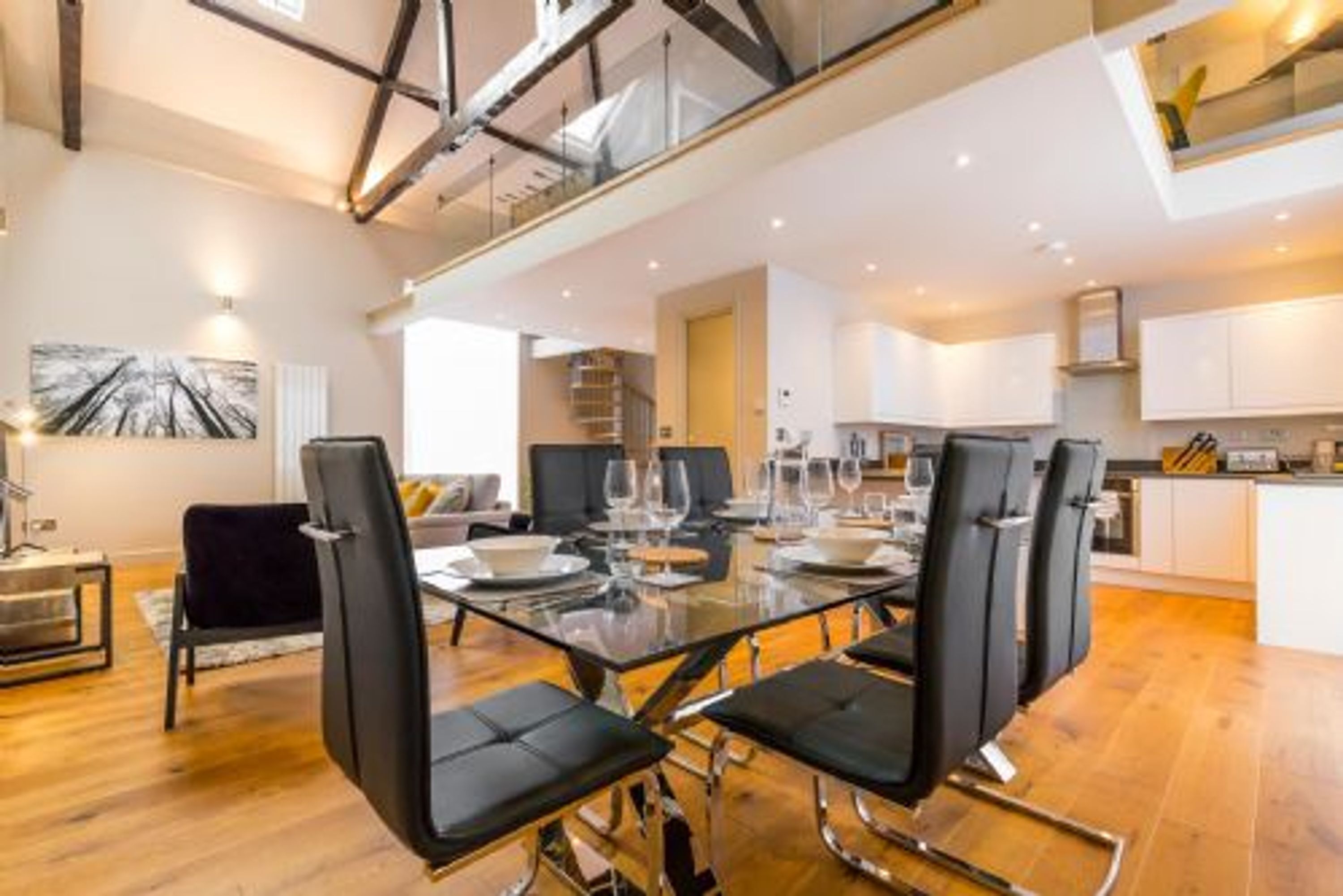 A modern dining area with a glass table, black chairs, and a kitchen in the background.