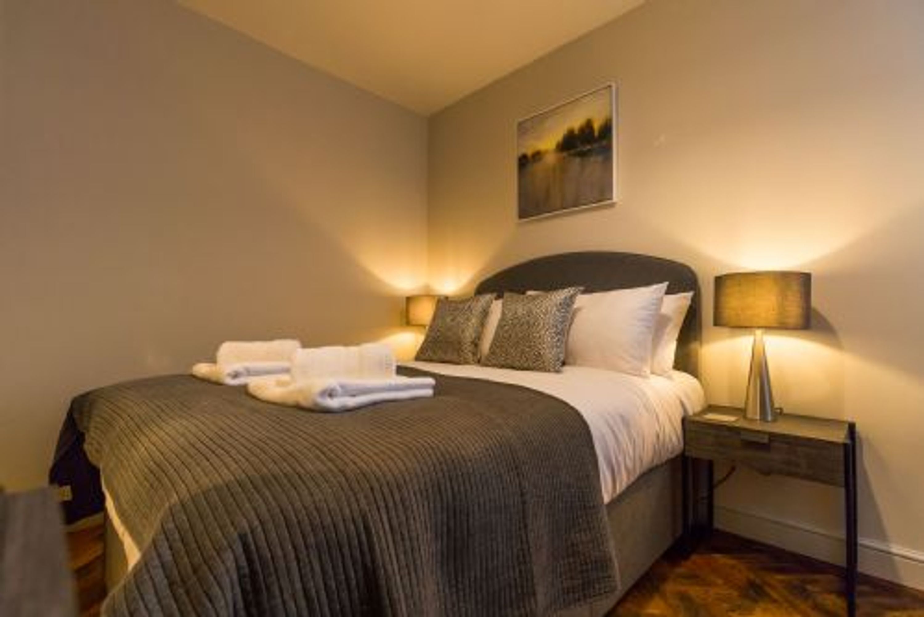 A neatly made bed with a dark headboard, a bedside lamp, and a painting on the wall.