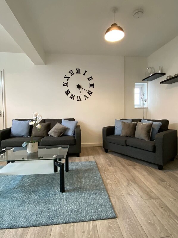 A living room with two dark sofas, a glass coffee table, a large wall clock, and a ceiling light. - Host Stay Waterfront (Northampton)