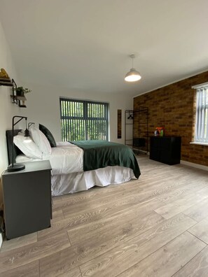A bedroom with a bed, a bedside table, a lamp, a window with blinds, a brick wall, and a floor lamp. - Host Stay Waterfront (Northampton)