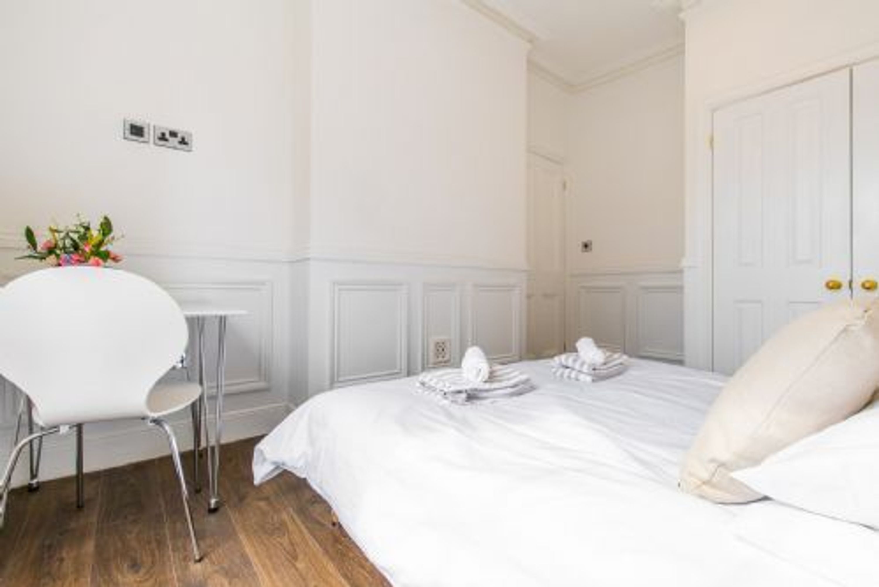 A hotel room with a bed, a white chair, and a small table with a vase of flowers.