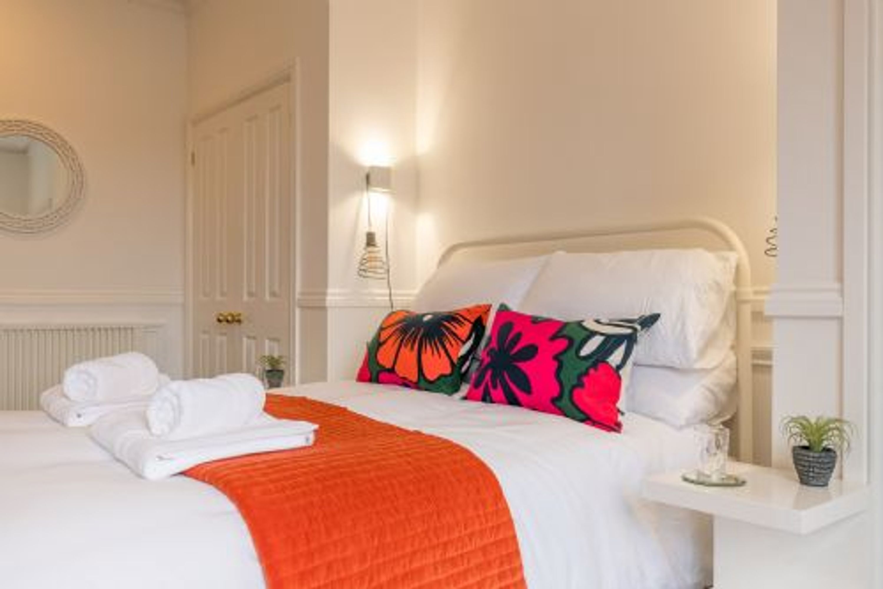 Cama bem arrumada com lençóis brancos e um cobertor laranja, mesa de cabeceira com uma planta e um espelho fixado na parede.