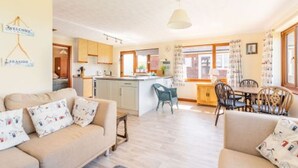 A cozy living room with a sofa and chairs, a kitchen area with cabinets and a dining table, and a welcoming sign on the wall. - Host Stay Breezy Bungalow (North Walsham)
