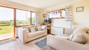 A spacious living room with a large window, a sofa, a television, and a lamp. - Host Stay Breezy Bungalow (North Walsham)
