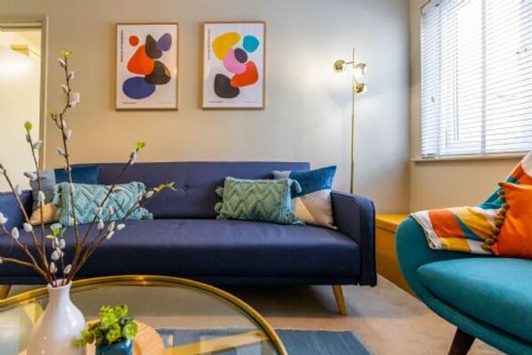A living room with a dark blue sofa, a round glass coffee table, and a colorful armchair. There are abstract art pieces on the wall and a vase with branches on the coffee table.