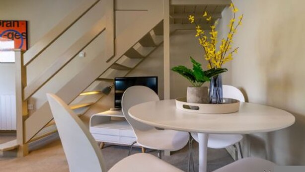 A modern interior with a round dining table, white chairs, a potted plant with yellow flowers, and a staircase. - Host Stay Corner Cottage (Norwich)