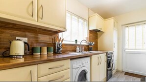 A compact kitchen with wooden countertops, a kettle, a washing machine, and a window with blinds. - Host Stay Corner Cottage (Norwich)