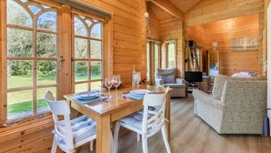 A wooden cabin interior with a dining area, a living room with a sofa, and large windows offering a view of the outdoors.