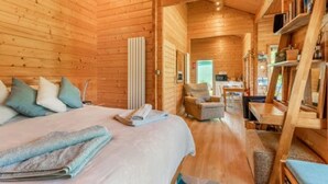 A wooden cabin interior with a bed, a chair, a table, and a bookshelf.