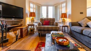 A cozy living room with a flat-screen TV, a plaid armchair, a grey sofa, and a coffee table with board games. - Host Stay St Joseph s Cottage (Sheringham)
