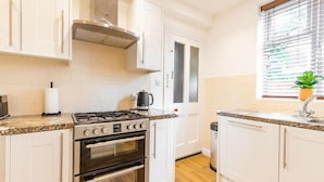 Une cuisine avec des meubles blancs, une cuisinière à gaz, un four, un évier et une fenêtre avec des stores.