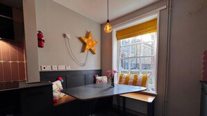 Une cuisine moderne avec un plan de travail foncé, un banc intégré agrémenté de coussins colorés, un luminaire en forme d’étoile et une fenêtre avec un store jaune.