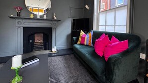 Un salon avec une cheminée, un canapé vert agrémenté de coussins colorés, un miroir rond et un téléviseur fixé au mur.