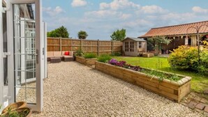 Un jardin avec un chemin de gravier, des jardinières en bois surélevées où sont plantées des fleurs, une clôture en bois et une maison avec un toit en tuiles.