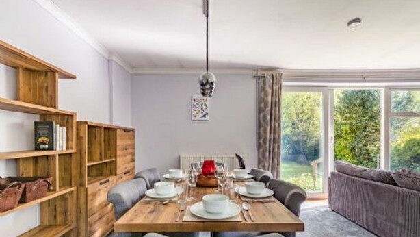 Un coin repas avec une table en bois dressée pour quatre personnes, entourée de chaises à revêtement gris. Une bibliothèque contenant divers objets se trouve à gauche.
