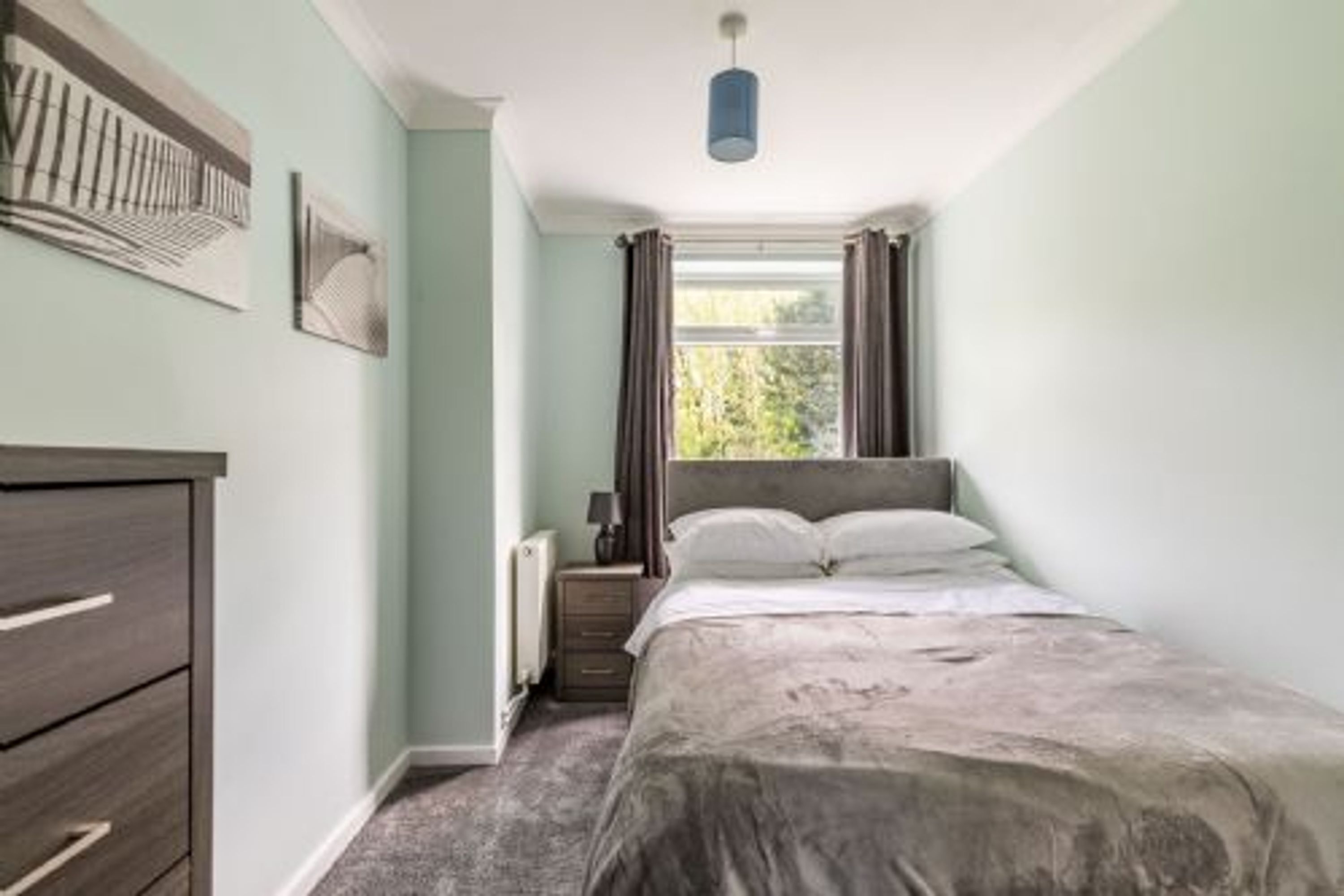 A bedroom with a bed, a dresser, a window with curtains, and a radiator.