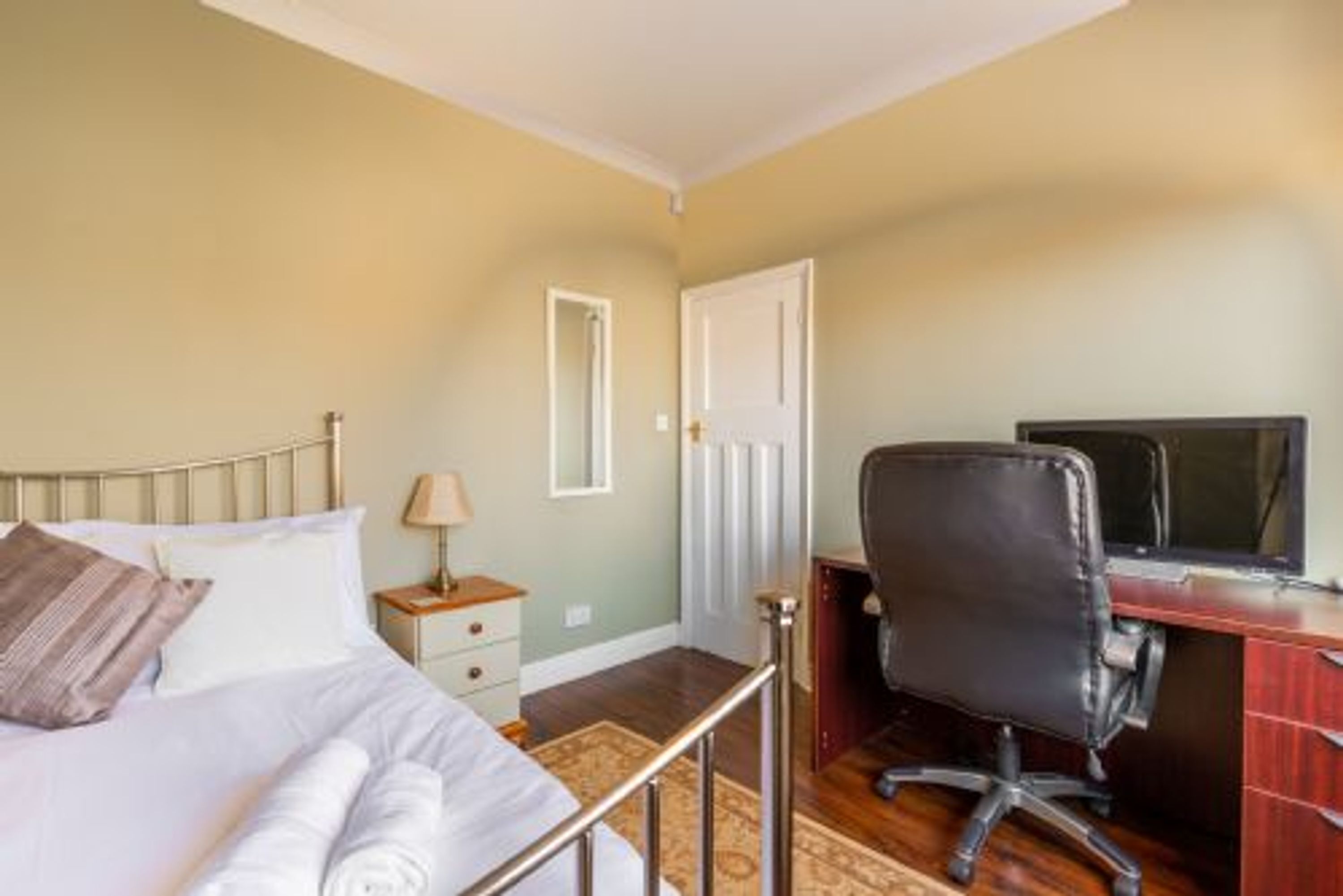 A hotel room with a bed, a desk with a computer, a chair, a lamp, and a door.