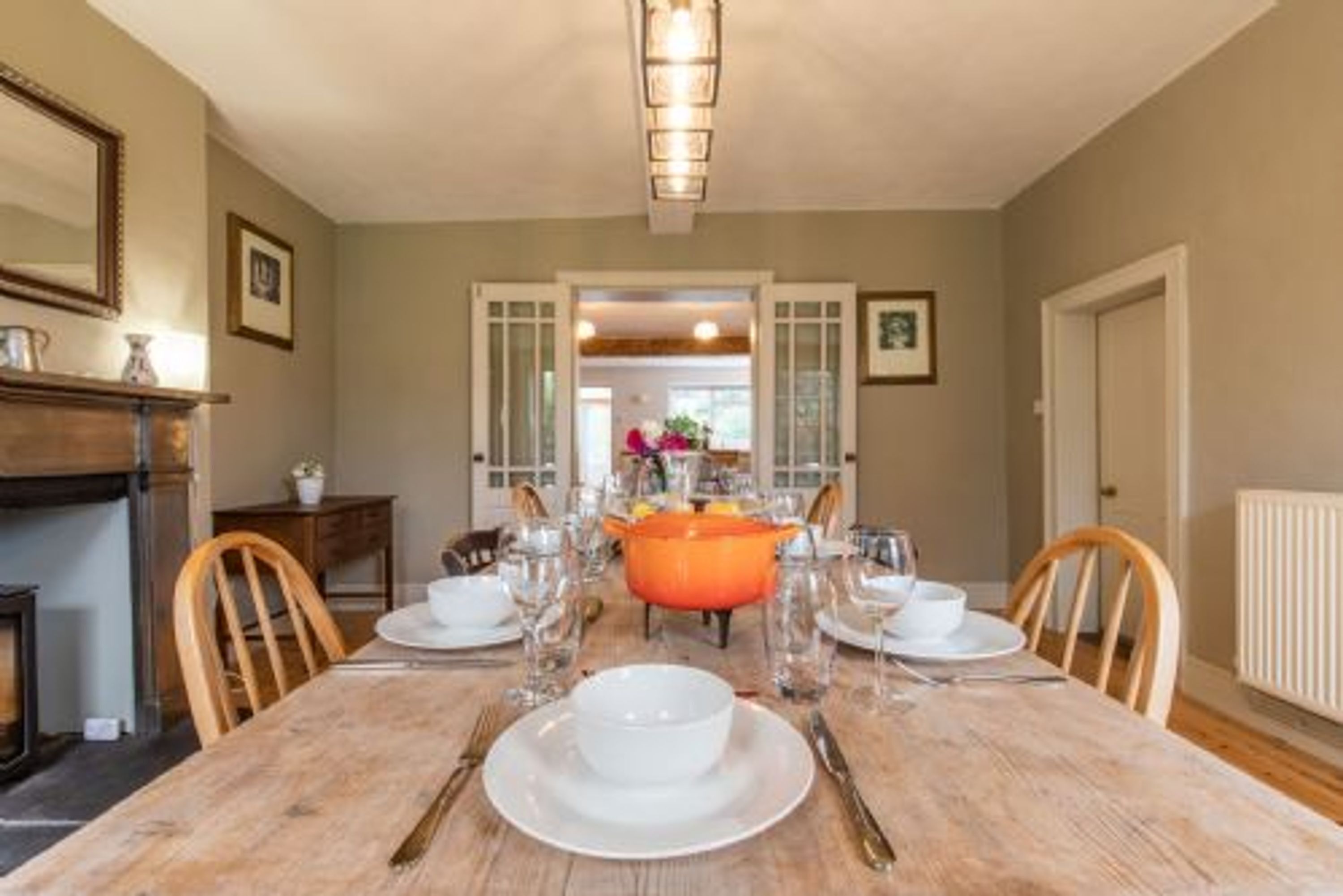 A dining room with a wooden table set for four, a large orange pot, and a fireplace in the background.