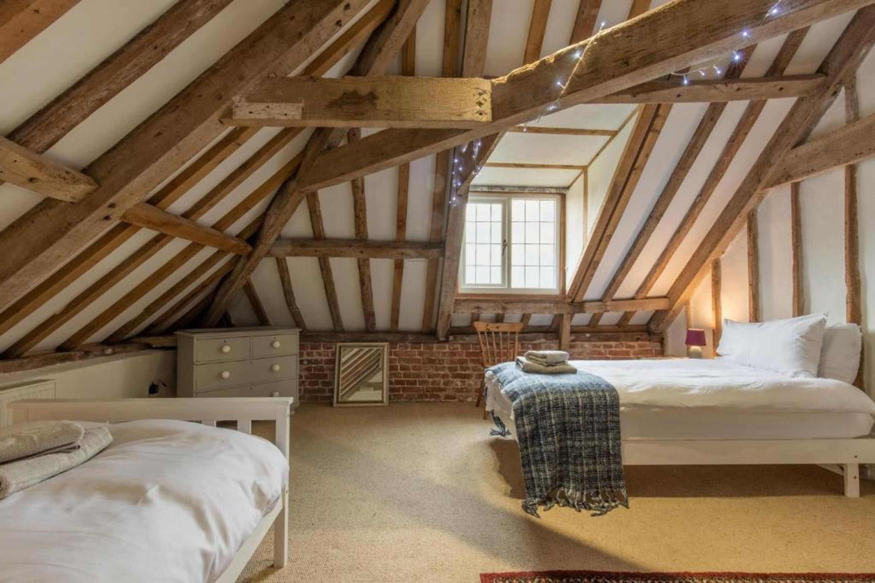 A bedroom with a sloped ceiling, wooden beams, a window, a bed, a dresser, and a mirror.
