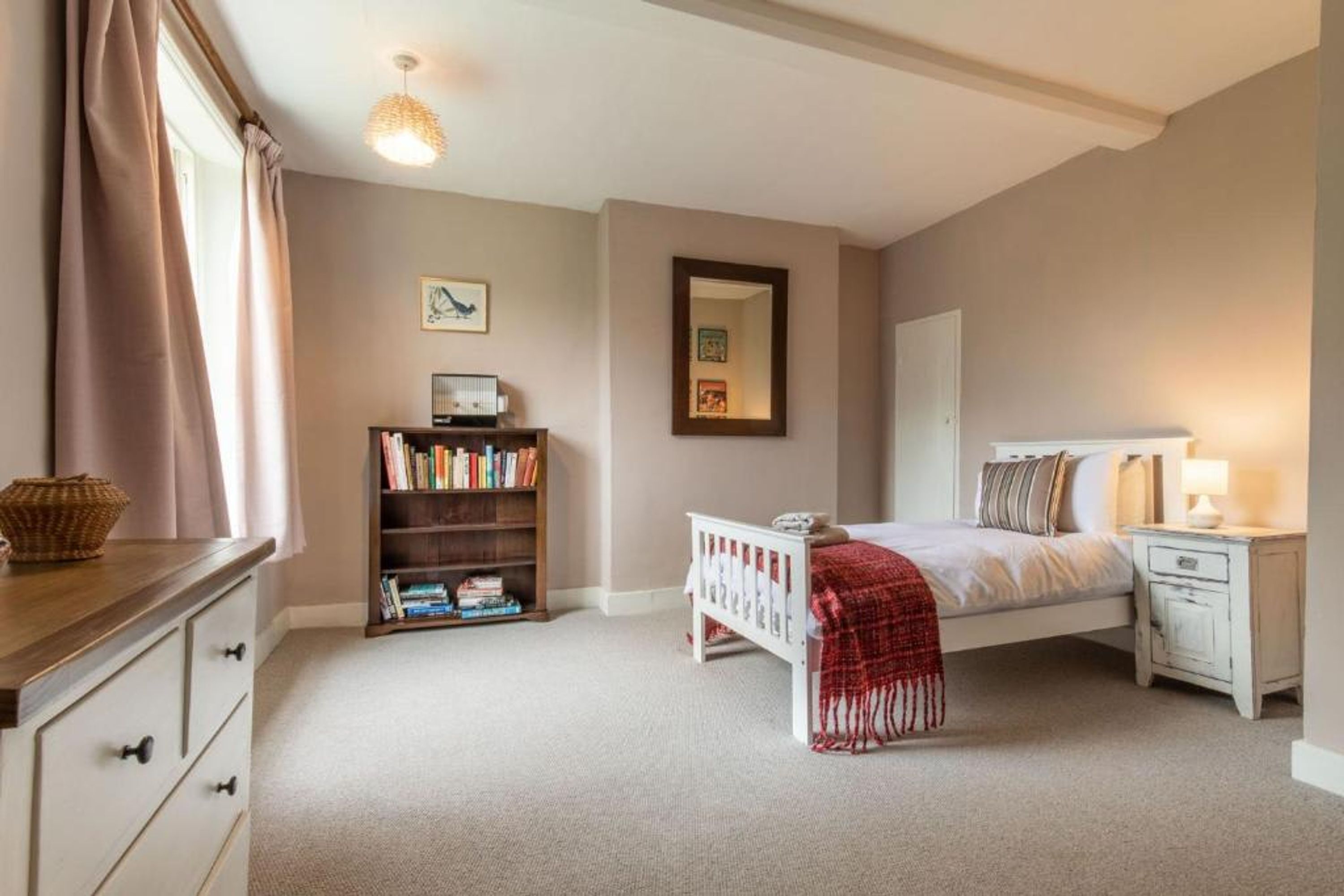 A bedroom with a bed, bedside table, lamp, bookshelf, and a window with curtains.