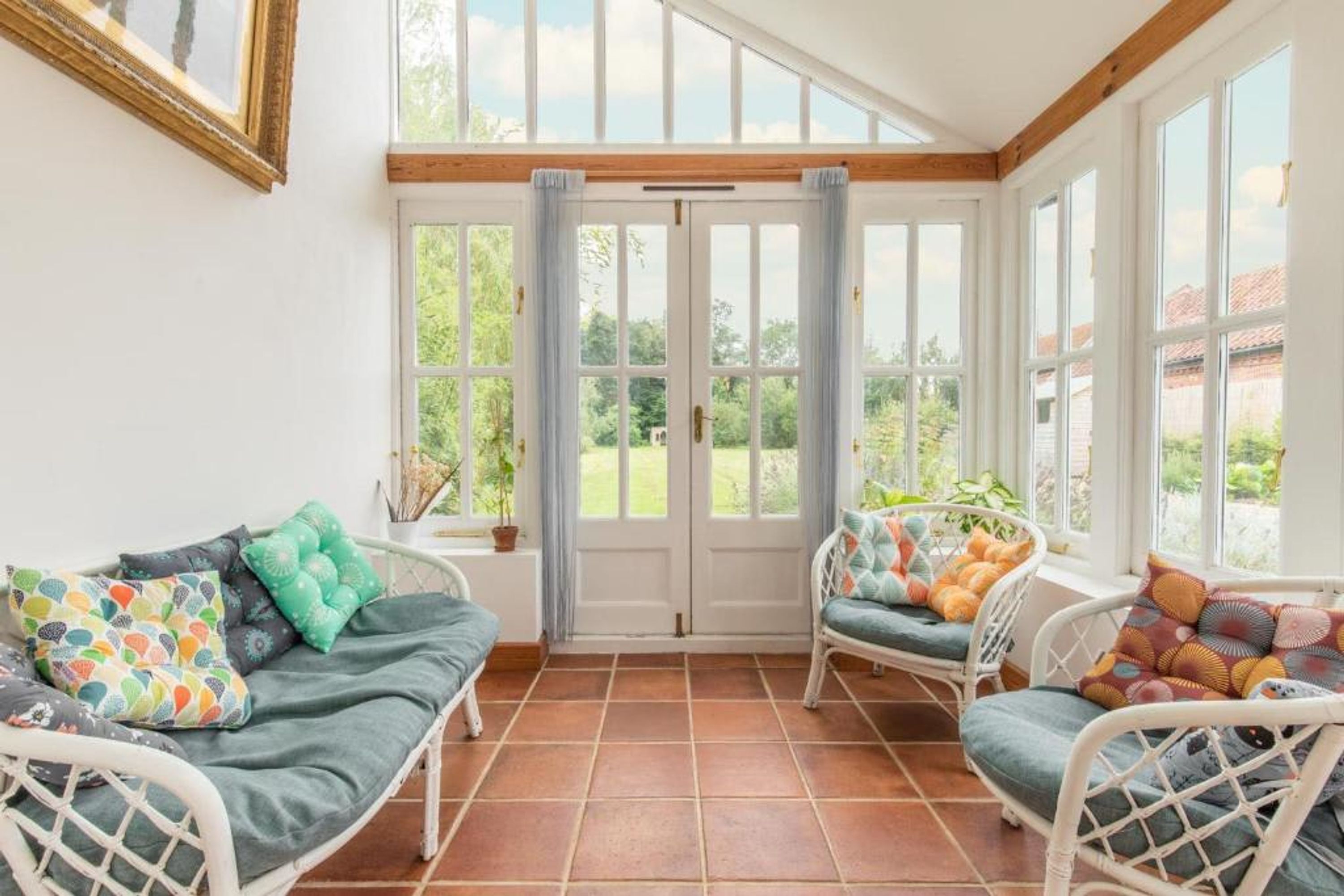 A sunroom with a view of greenery, white wicker furniture, and a tiled floor.