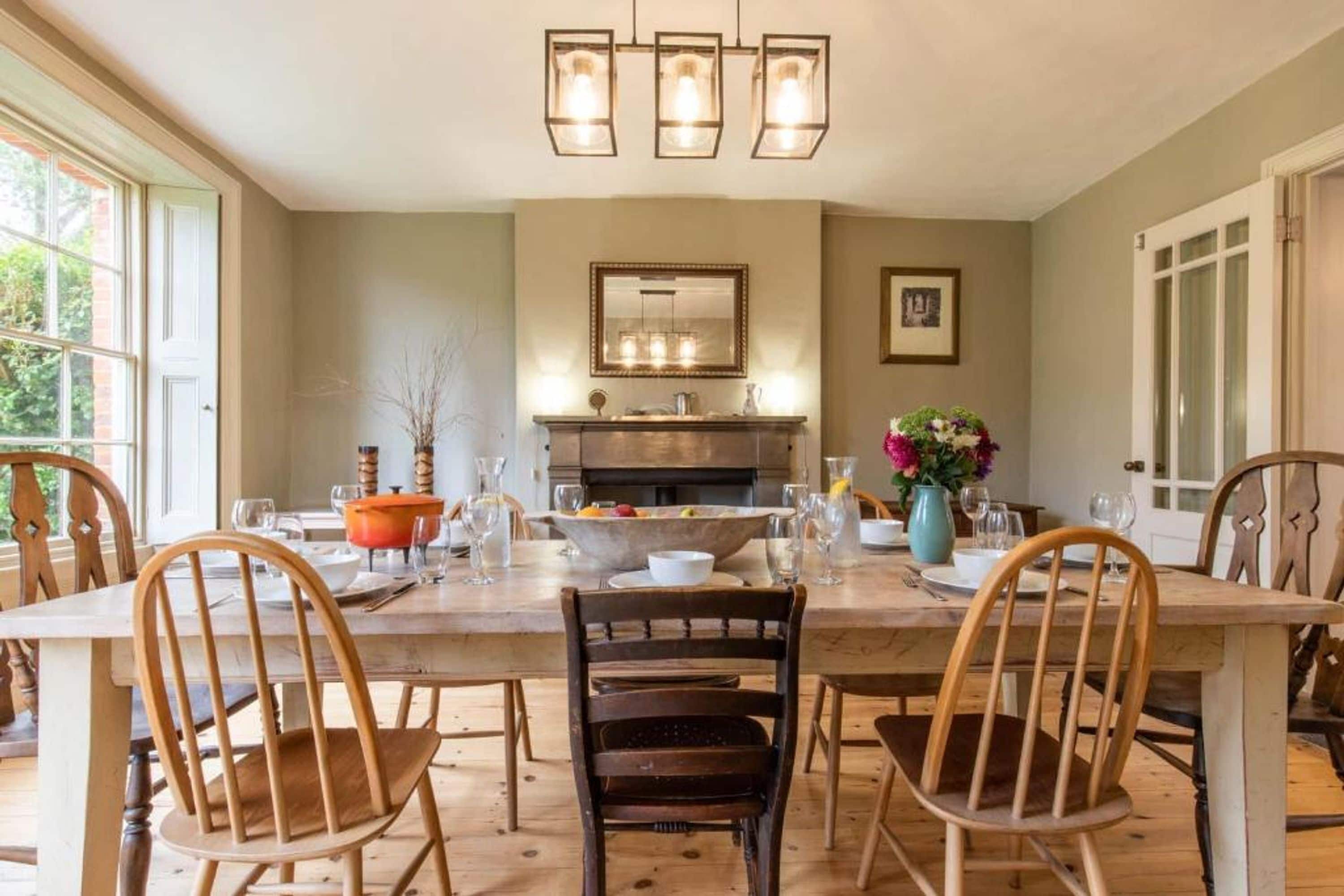 Un espace repas comprenant une longue table, des chaises, un lustre, un miroir et un vase rempli de fleurs.