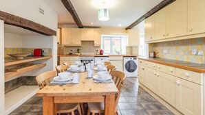 A kitchen with a dining table set for six, wooden cabinets, a washing machine, and a window providing natural light. - Host Stay 2 Ruscon Cottage (King's Lynn)