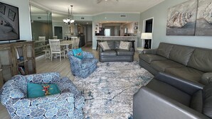 A spacious living room with a large gray sofa, a patterned blue sofa, and a gray armchair. There is a dining area with white chairs and a glass-top table. A kitchen is visible in the background with white cabinets and a black countertop.