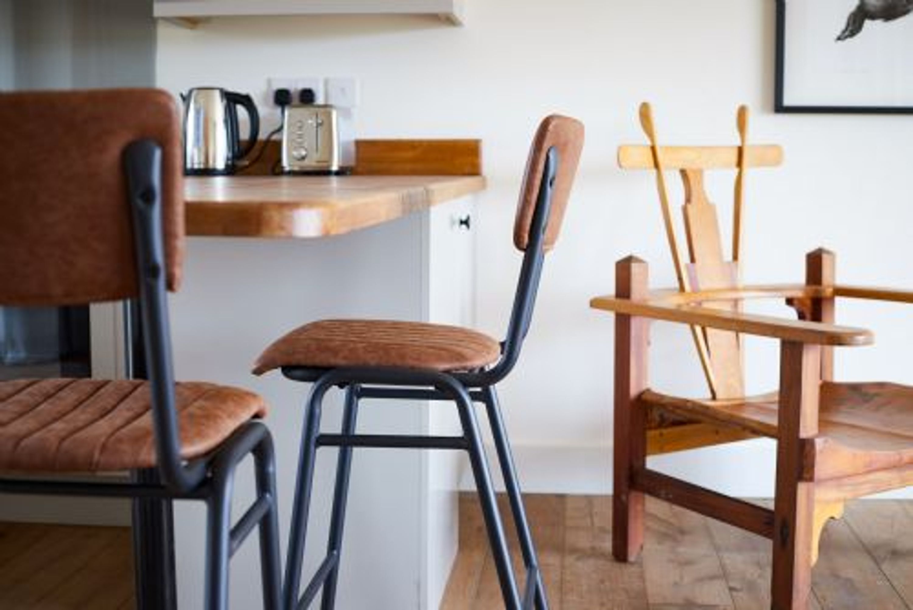A kitchen with a wooden countertop, a kettle, and a toaster. There are two chairs, one with a cushioned seat and backrest, and another with a wooden backrest and seat.