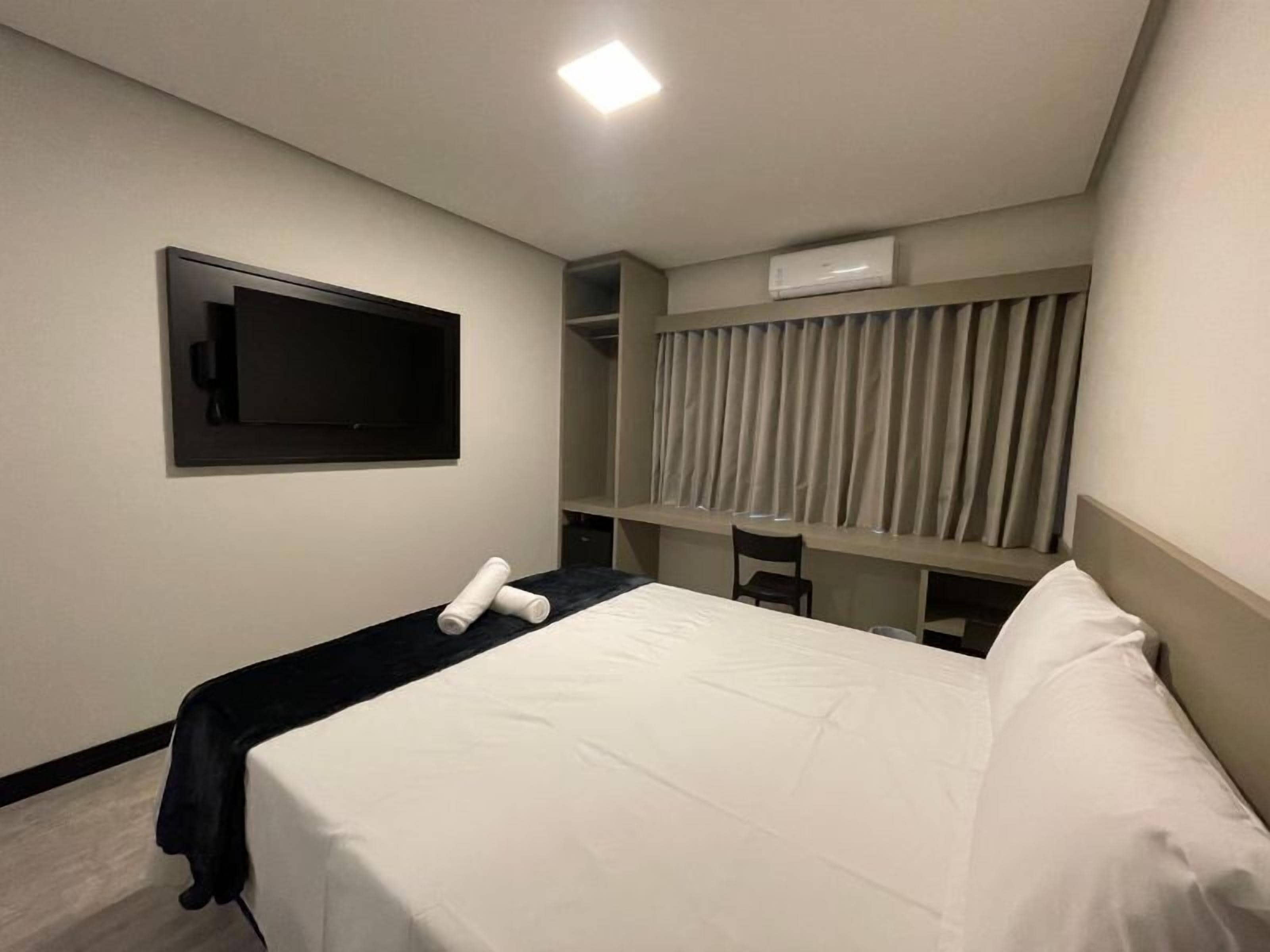 A hotel room with a bed, a TV mounted on the wall, a desk with a chair, and a wardrobe.