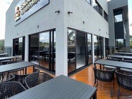 Uma área de refeições ao ar livre com mesas e cadeiras, grandes portas de vidro deslizantes e fachada de um edifício moderno.