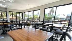 A spacious indoor dining area with large windows, wooden tables, and black chairs. - Escarpas Styles Hotel (Capitólio)