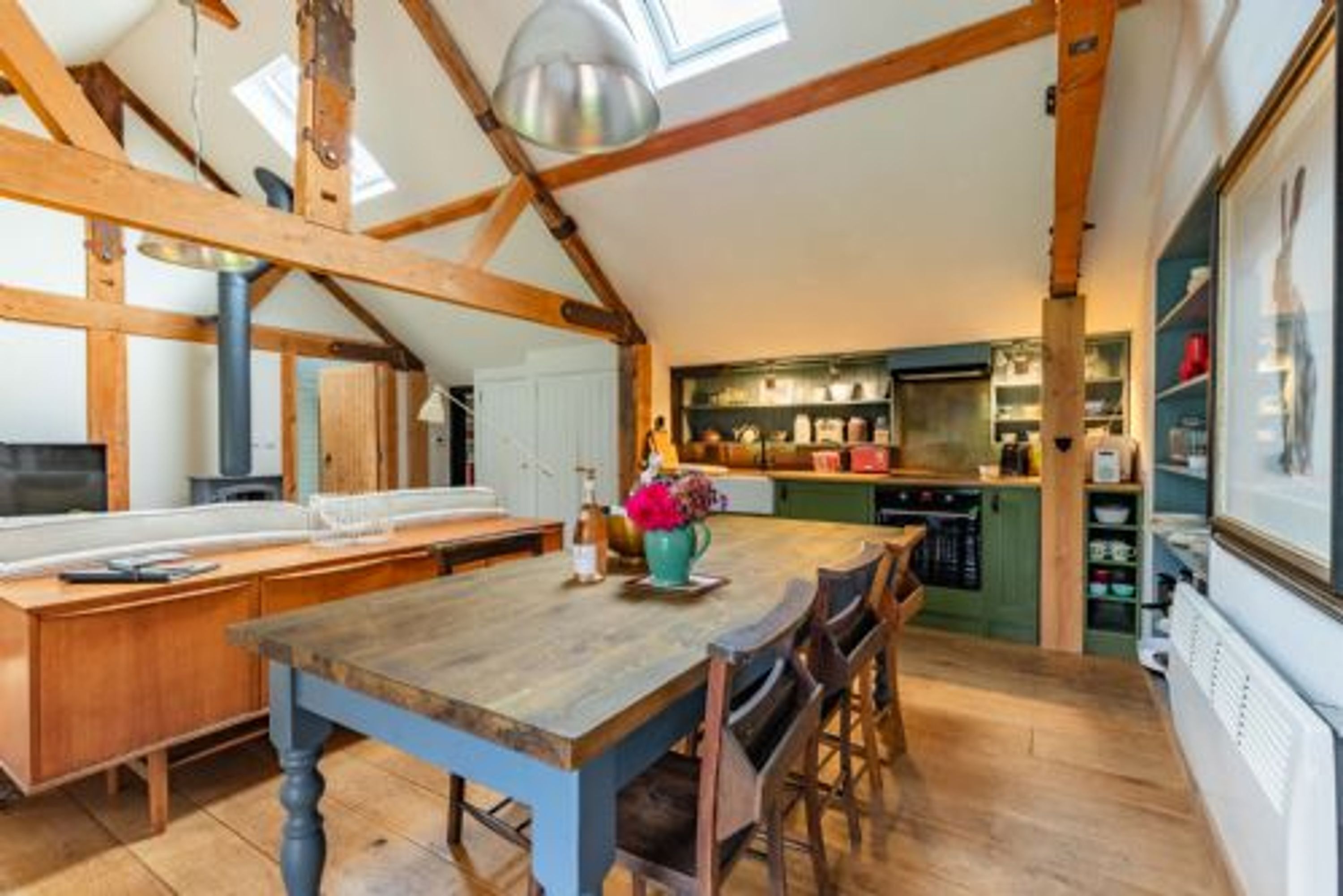 A rustic kitchen with wooden beams, a large island table, and a view of the living area.