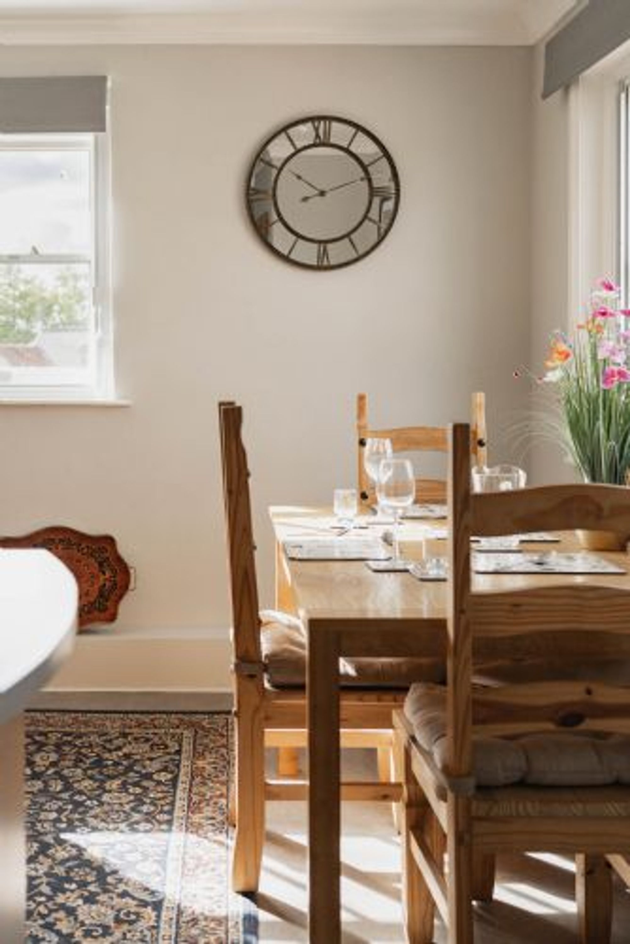A dining area with a wooden table, chairs, a wall clock, and a window with a view of the outdoors.