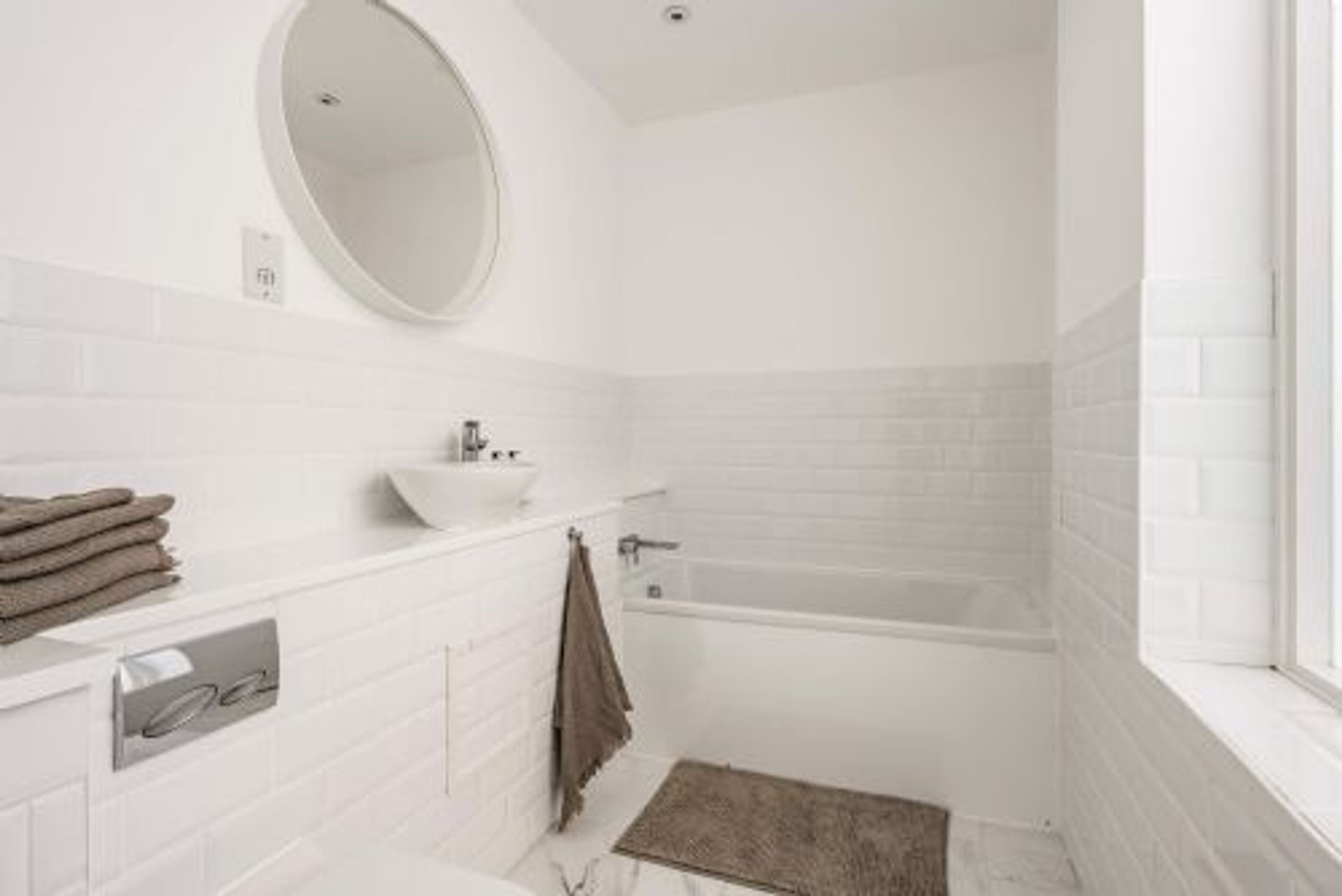 A modern bathroom with a white tiled wall, a round mirror, a sink, and a bathtub.