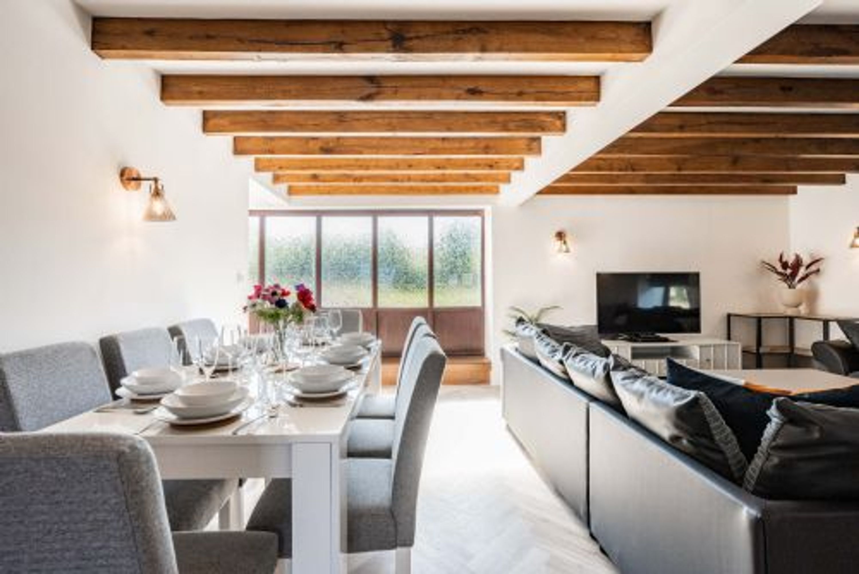 A modern dining area with a long table set for a meal, a sofa, and a television in the background.
