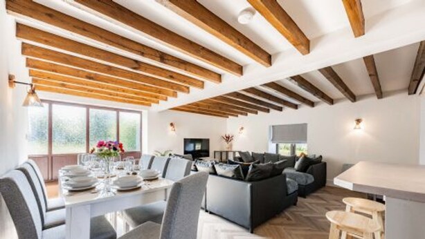 Un intérieur moderne avec des poutres apparentes, un coin repas avec une table blanche et des chaises grises, un canapé gris et un espace cuisine doté d’un plan de travail blanc et de tabourets en bois.