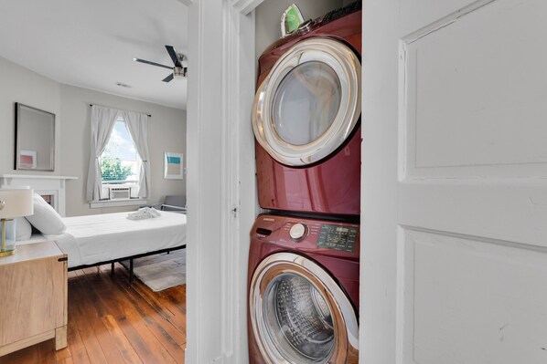 A bedroom with a bed, a nightstand, a lamp, a window with curtains, and a washing machine. - Spacious Updated Home Ideal For Groups W/ Parking by Lucky Savannah (Savannah)