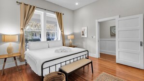 Un dormitorio con una cama, lámparas de noche, un banco, una ventana con cortinas y una puerta.