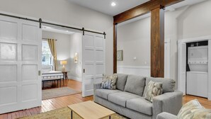 Una sala de estar moderna con un sofá gris, vigas de madera, una puerta blanca y una zona de cocina con un refrigerador blanco.