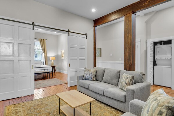 A modern living room with a grey sofa, wooden beams, a white door, and a kitchen area with a white refrigerator.